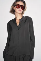 Camisa negra de lyocell con cuello redondo, manga larga con puños y bajo asimétrico con aberturas laterales. Cierre frontal con botones.