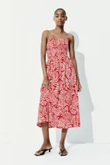 Vestido midi de breteles finos con estampado de hojas en color blanco sobre fondo rojo. El cuerpo es de punto smock, tiene escote cuadrado, costura fruncida en la cintura y falda con vuelo.