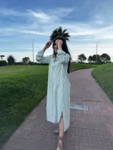 Vestido camisero midi a rayas verticales color verde y blanco, con cuello camisero, mangas largas con puños abotonados, cierre frontal con botones y aberturas laterales en el ruedo.