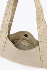 Bolso de hombro color beige, confeccionado en serraje, con tachas metálicas doradas en el cuerpo y las asas. Cuenta con un bolsillo interior y cierre mediante botón imantado.