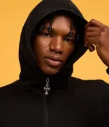 Saco tipo hoodie con cierre frontal de cremallera, color negro, corte boxy y capucha. Presenta estampa de letras en blanco en ambas mangas y un colgante de cruz metálico en el tirador de la cremallera principal.