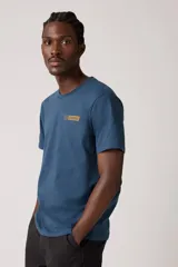 Remera de manga corta, cuello redondo, color azul oscuro, con un pequeño parche gráfico rectangular con texto en tono mostaza en el pecho.