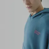 Canguro azul petróleo con capucha y bolsillo delantero, con logo 'Human Being' bordado en rosa en el pecho.