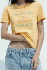 Camiseta color amarillo con efecto lavado, cuello redondo y mangas cortas. Estampado frontal con un oso de los Care Bears™ y la frase "free hugs".