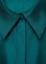 Camisa verde esmeralda de corte recto y tejido satinado, con cierre frontal de botones ocultos, cuello camisero, mangas largas con puños abotonados y bajo asimétrico.
