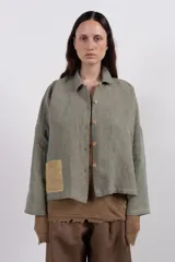 Camisa amplia de lino en tono verde desgastado, con textura visual y caída liviana. Destaca por su parche tejido a mano en contraste, que aporta un detalle artesanal y único.