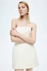 Vestido mini strapless color crudo, confeccionado en sarga de mezcla de algodón con costuras visibles y cuerpo ceñido con varillas. Presenta cintura interior de grosgrain con cierre ajustable de corchete, cierre oculto con cierre de corchete en la espalda y cadera sobredimensionada. Forrado.