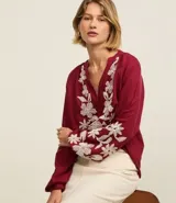 Blusa color borgoña de batista con cuello en V y botones. Presenta bordados florales blancos en el frente y las mangas largas con puños elásticos.