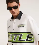 Camisa blanca oversized de tricolina con cuello negro contrastante, mangas cortas y estampado con la palabra "Hustle" en verde y negro.