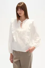 Blusa blanca de calce holgado con cuello en V y lazo frontal. Presenta detalles de volados en el pecho y cuello, y mangas largas con puños elásticos.