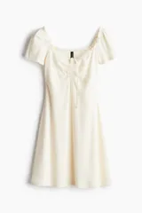 Vestido corto color crema, de punto acanalado, con escote corazón ribeteado en encaje y cordón para ajustar en el centro del busto. Modelo de manga corta con cuerpo entallado y falda con vuelo.