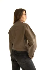 Chaqueta corta de cuero marrón con cuello mao, cierre frontal con botones a presión, dos bolsillos de parche con solapa en el pecho y mangas largas abullonadas con puños elásticos.