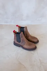 Botas Chelsea de cuero color marrón, con elásticos laterales negros, tirador trasero rojo y suela de goma negra con costuras rojas.