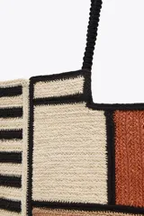 Bolso tipo tote de yute con diseño de bloques de color y rayas horizontales en negro y beige. Asas de hombro negras.