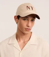 Gorra de sarga color beige con bordado de las letras "NY" en marrón. Tiene visera curva y detalle de cuerda trenzada en la base de la gorra.