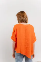 Blusa color naranja confeccionada en 100% lino, con escote en V, mangas cortas con vuelta y silueta amplia. Presenta terminación asimétrica y fila de botones decorativos en uno de los hombros.