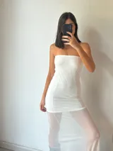 Vestido largo blanco, estilo tubo, sin tirantes (strapless). La parte superior es opaca y ajustada, mientras que la falda es de tela transparente que deja ver las piernas.
