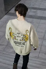 Buzo color arena de corte oversized, con cuello redondo y puños y cintura acanalados. Estampado en la espalda con dos tigres enfrentados y texto "Pray x Mercy" y "Against All Since XIX".
