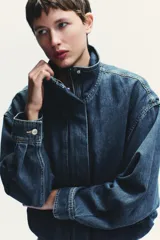Chaqueta corta de denim azul con cuello alto, manga larga con hombros caídos, bolsillos laterales, bajo con elástico y cierre frontal con cremallera y botones ocultos.