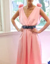 Vestido largo color rosa pálido, con escote en V cruzado, sin mangas y cinturón negro con apliques de flores. Presenta detalle de volados en los hombros y corte evasé.