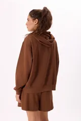 Conjunto deportivo color marrón, compuesto por un buzo con capucha, cierre frontal, bolsillos y terminaciones metálicas, y un short con cintura elástica ajustable con cordón.