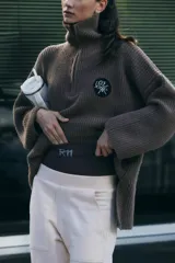 Sweater marrón de punto con cuello polo, cierre de cremallera parcial y parche bordado en el pecho.