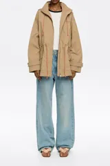 Gabardina corta color beige, de tejido técnico, corte línea A, cuello alto con capucha ajustable con cordones, manga larga recta con puño de presilla y botón a presión. Cierre frontal con cremallera metálica, cintura ajustable con cordón personalizado y logo estampado en el exterior.