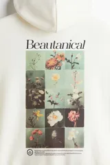Buzo canguro color crema con capucha cruzada de doble capa, hombros caídos, bolsillo tipo canguro, puños y bajo acanalados. Estampado frontal con flores y texto "Beautanical". Corte holgado.