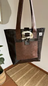 Bolso tote de ecocuero marrón oscuro con textura cocodrilo y un parche frontal de cuero liso marrón claro. Posee asas de hombro de cuero marrón y un broche decorativo blanco en el frente.