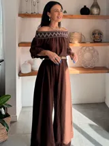 Enterito largo color blanco, con escote Bardot fruncido y mangas cortas abullonadas. La cintura es ajustable con un cinturón ancho de rafia con hebilla redonda. El pantalón es ancho y largo.