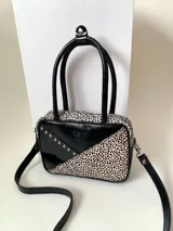 Cartera rígida negra de cuero con tachas plateadas y tres combinaciones de cuero diferentes que aportan textura. Tiene herrajes y cierre metálico plateado marca YKK. Cuenta con un asa fina para llevar al hombro o en la mano y una correa larga extra regulable.