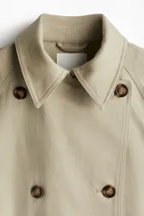 Gabardina midi color beige de algodón, con doble botonadura, cuello con solapas, cierre de corchete en la parte superior, solapa de tormenta en la espalda, bolsillos laterales con solapa, cinturón extraíble con hebilla rectangular, mangas raglán con trabilla ajustable en los puños y abertura trasera.