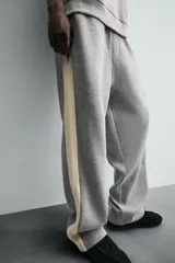 Pantalón de jogging gris con corte relaxed fit, cintura elástica ajustable con cordón interior, bolsillos laterales y traseros de vivo, y banda lateral color crema.
