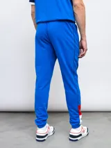 Pantalón de jogging azul marino con puños ajustados, cintura elástica con cordón y bolsillos laterales. Presenta una franja blanca con detalles en rojo a lo largo de la pierna, el escudo del Club Nacional de Football y el logo de Umbro.