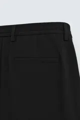 Pantalón negro de corte ancho con cintura elástica, pliegues frontales, bolsillos laterales y traseros de vivo. Tiene detalle de hebilla metálica en la trabilla y cierre frontal con cremallera y botón.