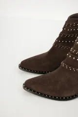 Bota corta de cuero color marrón oscuro (chocolate) con punta afilada y taco bajo. Presenta múltiples tiras decorativas con hebillas y tachas plateadas alrededor del empeine y el tobillo, además de tachas en el borde de la suela.