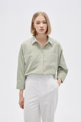 Camisa color verde claro, de corte holgado, con cuello clásico, cierre frontal con botones, mangas 3/4 y dos bolsillos de parche en el pecho.