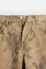 Pantalón de jean color beige con estampado abstracto floral en tonos marrones, de corte amplio, con bolsillos delanteros y traseros, y detalle de tira multifunción en la pierna. Cierre frontal con cremallera y botón.