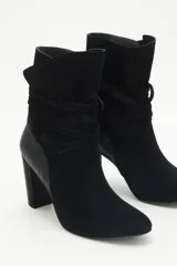 Botas de caña media de gamuza negra, con taco cuadrado y punta fina. Presenta tiras terminadas con borlas que se cruzan y atan alrededor de la caña.