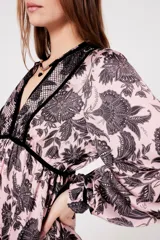 Vestido largo rosado con estampado floral negro, escote en V con ribete negro, mangas largas abullonadas y falda escalonada con volantes.