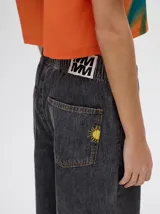 Mono largo de jean celeste con corte holgado, cintura elastizada con cordón ajustable, sin mangas, cuello camisero y cierre frontal con botones. Presenta bordados de motivos playeros en el pecho.