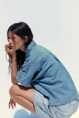 Camisa de denim celeste con cuello solapa, manga corta, bolsillo de parche en el pecho, bajo con aberturas laterales y cierre frontal con botones.