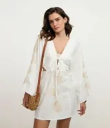 Vestido corto de playa confeccionado en voil de algodón BCI color blanco, con escote en V con lazo, cutouts laterales, mangas largas y bordados de caracoles de mar.