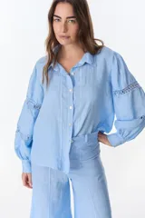 Camisa celeste de lino con cuello clásico, abotonada al frente y mangas largas con detalle de puntilla en los hombros.