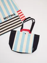 Bolso tote grande con estampado a rayas verticales celestes y blancas, laterales negros y asas cortas negras con refuerzo rojo. Tiene cierre superior y está confeccionado en plástico con doble tela.