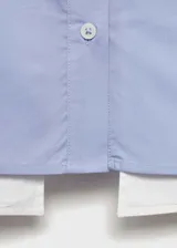 Camisa celeste de popelina de algodón con diseño crop, cuello camisero, cierre frontal con botones, dos bolsillos de solapa en la parte delantera, manga larga con puños abotonados y terminaciones en contraste blanco.