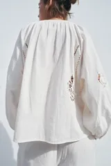 Camisa color crudo confeccionada en mezcla de algodón y lino, con cuello redondo y escote pico ajustable con lazadas. Tiene manga larga abullonada y detalle de bordados perforados a tono.