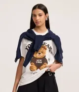 Remera blanca de algodón con cuello redondo y manga corta. Presenta un estampado central de un oso de peluche marrón vistiendo un buzo negro con el texto "NEW YORK" en blanco.