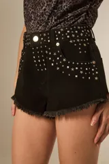 Short de jean negro, tiro medio, ajustado al cuerpo, con roturas y tachas plateadas en forma de estrellas y cuadrados en la parte delantera. El ruedo es deshilachado.