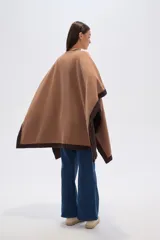 Poncho de tejido grueso color camel, con bordes y mangas en color marrón oscuro. Cierre frontal con broche rectangular negro.